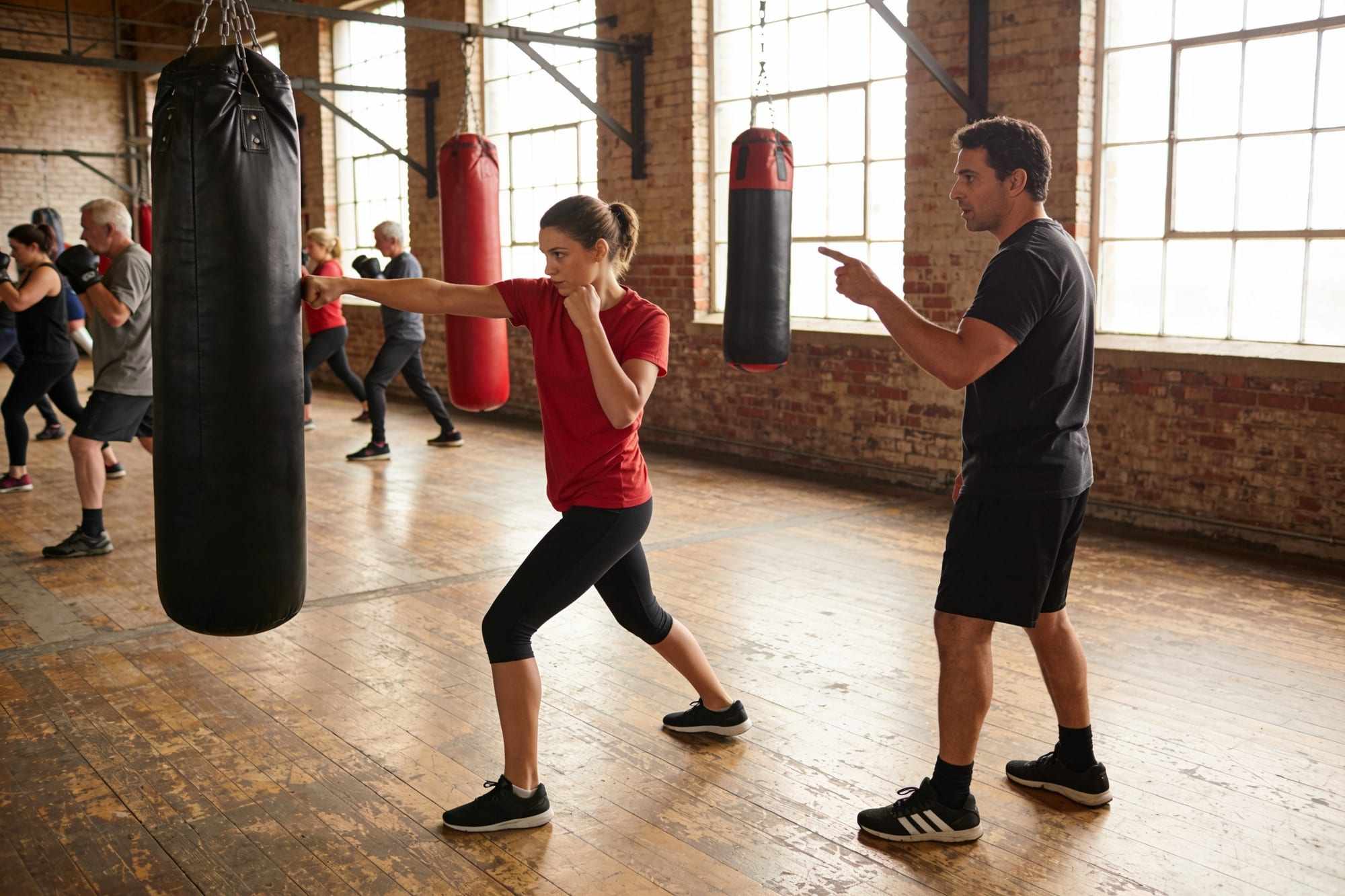 A beginner in a red T-shirt throwing a jab at a heavy bag while a coach watches her feet